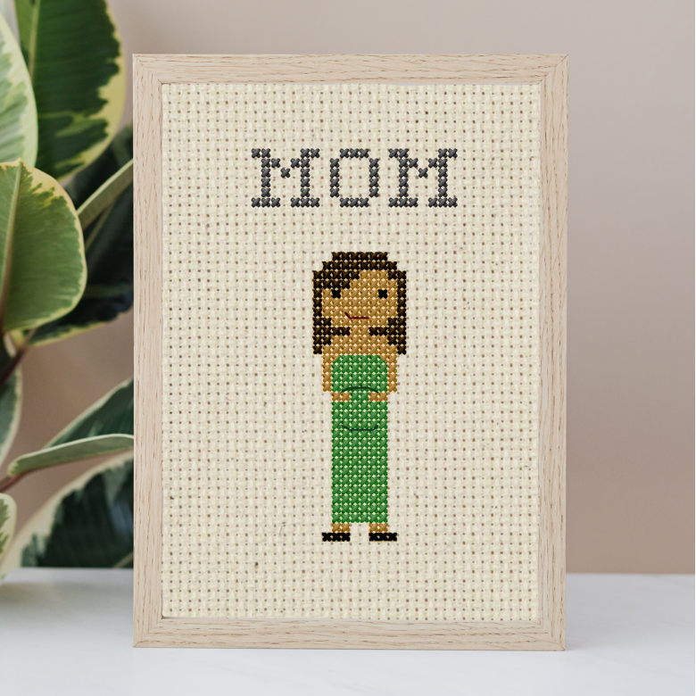 Framed cross-stitched artwork of a woman labeled 'MOM' with a plant in the background