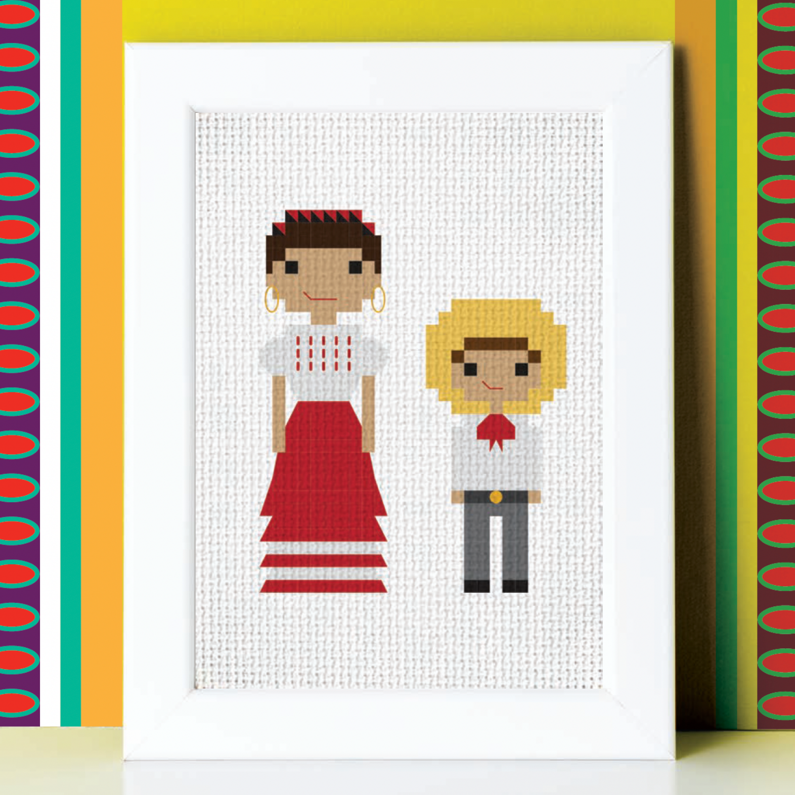 Framed cross-stitch art of two people against a colorful geometric-patterned wall.