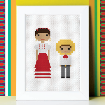 Framed cross-stitch art of two people against a colorful geometric-patterned wall.