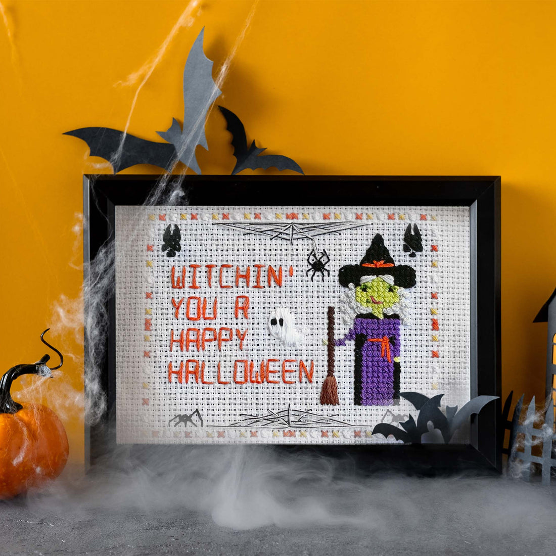 Framed Halloween-themed cross-stitch with decorative pumpkins and a small house on a yellow background.